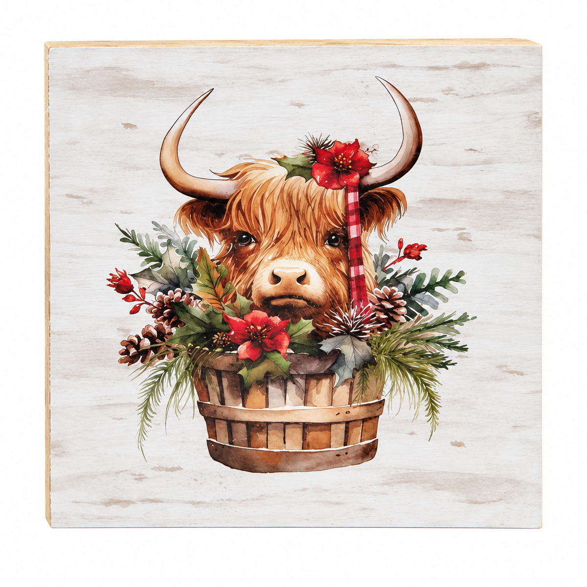 A square decorative wall plaque featuring a painted Highland cow with curved horns emerging from a wooden basket filled with red flowers, green foliage, pinecones, and a red-and-white striped ribbon, set against a light weathered wood background.
