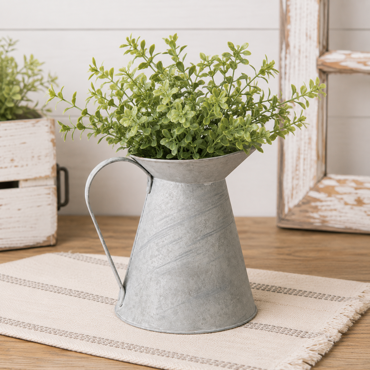 Galvanized metal pitcher planter filled with green foliage, displayed on a woven placemat on a wooden table with rustic home decor in the background.