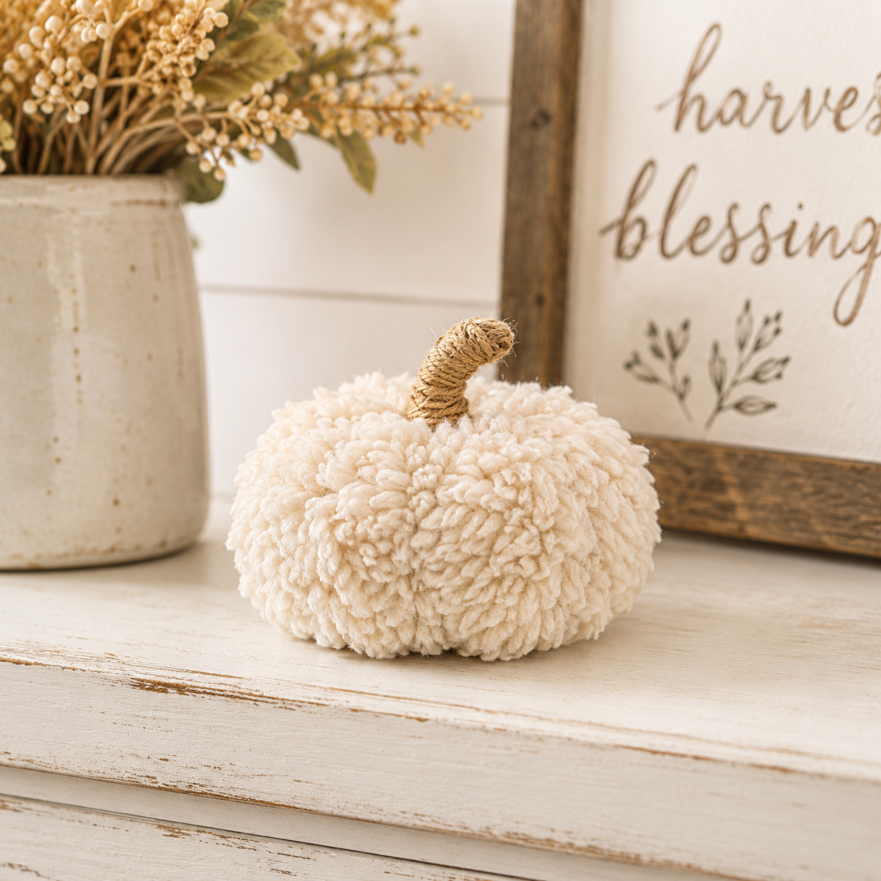 A small decorative faux pumpkin in a textured cream bouclé fabric with a twisted natural jute stem, styled on a distressed white wooden surface next to a ceramic vase with dried flowers and a framed sign.