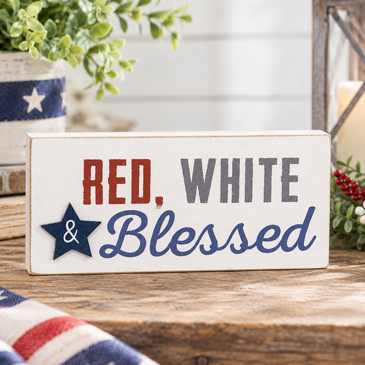 Distressed white wooden “Red, White & Blessed” block sign with red and gray lettering, blue script, and a dimensional navy star with white ampersand, displayed on a rustic wood tabletop in bright farmhouse-style decor with leafy greenery, red and white berry sprigs, a candle lantern, and softly arranged patriotic red, white, and blue accents.