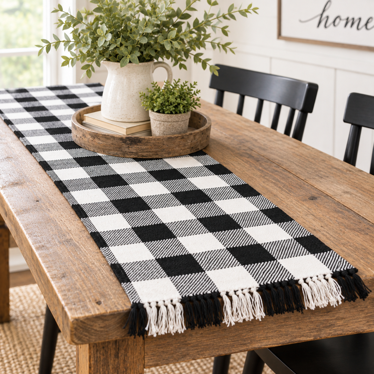 A black and white buffalo plaid table runner with fringe tassels, styled on a rustic wooden dining table with a wooden tray, books, and potted greenery.