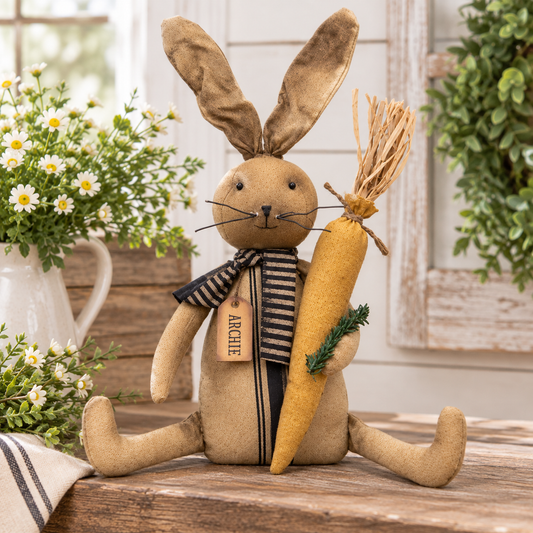 A rustic fabric bunny doll with long tan ears, a striped navy and beige scarf, and a hanging tag that reads "ARCHIE," sitting on a wooden surface and holding a large felt carrot with a straw top. The scene is styled with flowers, greenery, and a white shiplap wall background.