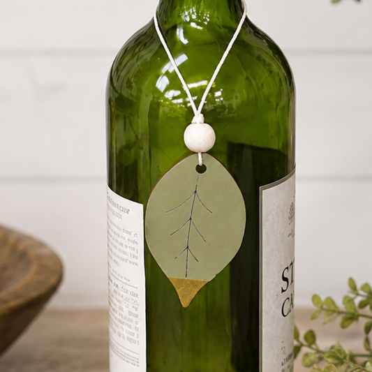 A green glass wine bottle with a purple neck wrap and a hanging leaf-shaped tag, styled on a wooden table with books, a candle, and a ceramic vase in the background.