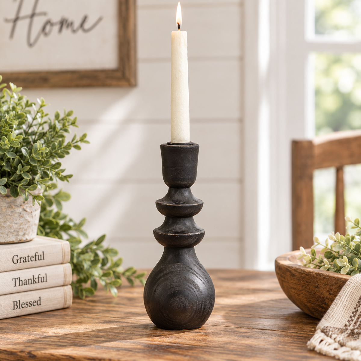 A black wooden tiered candlestick holder with a lit white taper candle, displayed on a rustic wooden table in a bright, sunlit room with home decor props.
