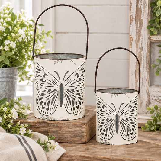 Two distressed white metal lanterns with black butterfly designs and dark metal handles, displayed on a rustic wooden surface with potted plants and a white paneled wall in the background.