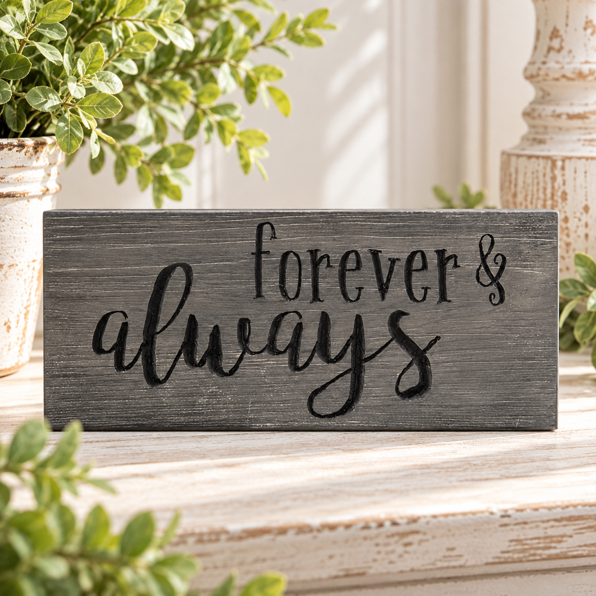 A rectangular dark gray wooden sign with the words 'forever & always' in black script, displayed on a distressed white wooden surface with potted green plants in the background.