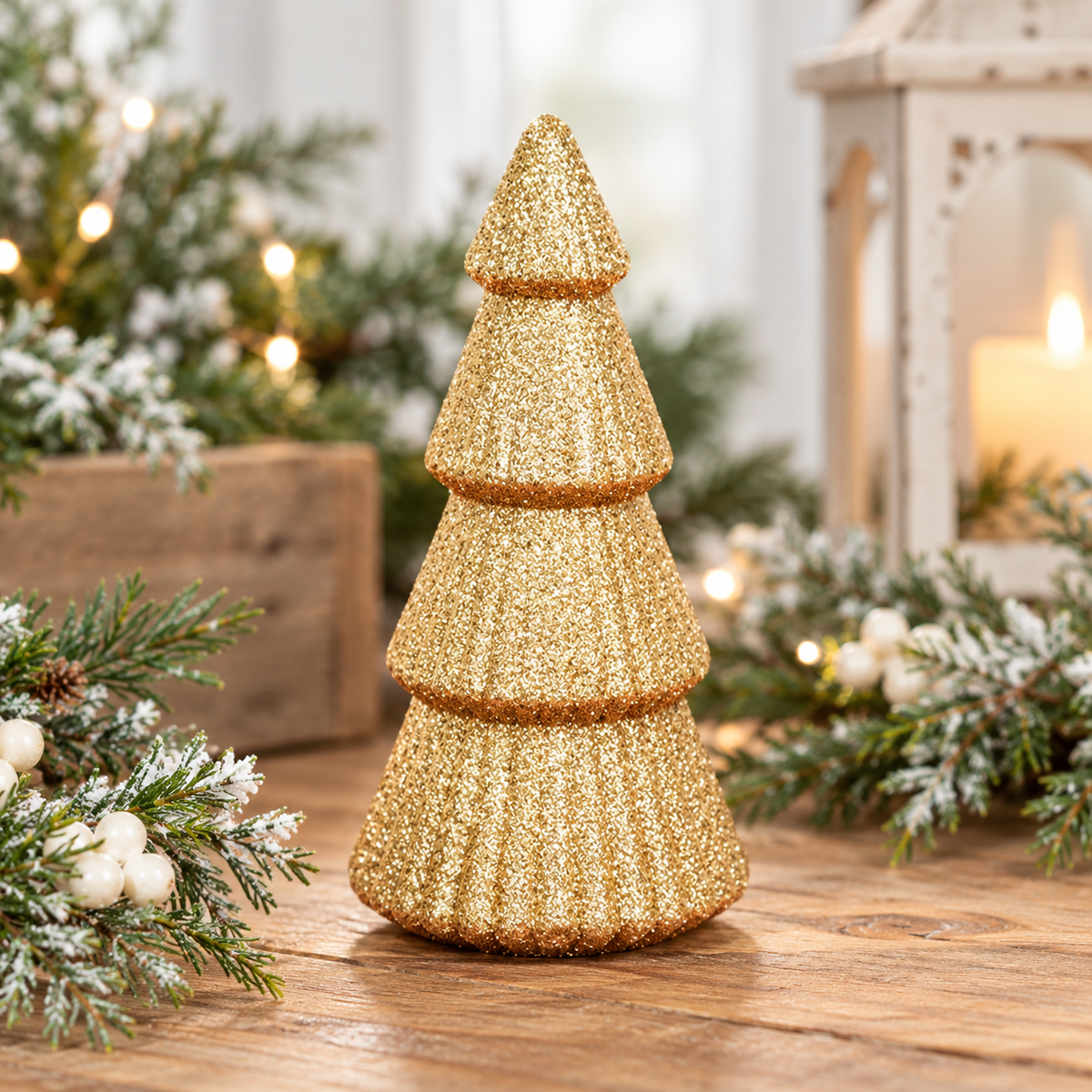 A small tiered Christmas tree decoration covered in textured gold glitter, standing on a wooden surface with festive greenery and a warm lantern in the softly blurred background.