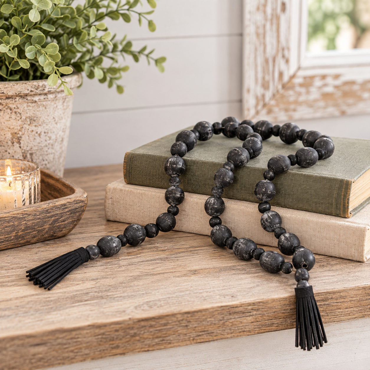 A black beaded garland with two black tassels, draped over two stacked vintage-style books on a wooden table, with a potted plant and a lit candle in the background.