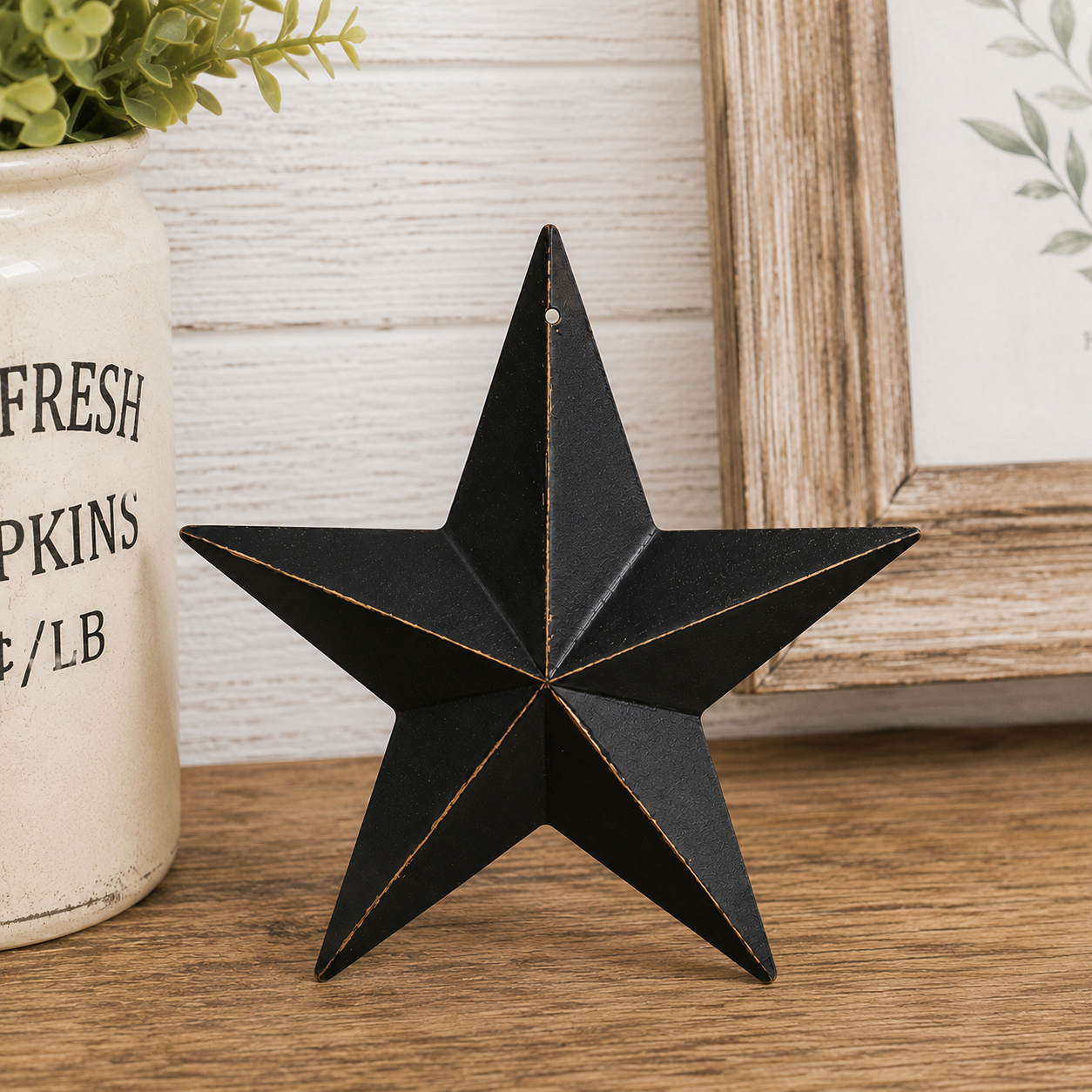 A black metal five-point star decoration with gold-toned edge trim, displayed on a wooden tabletop next to a cream-colored ceramic jar and a rustic framed print.
