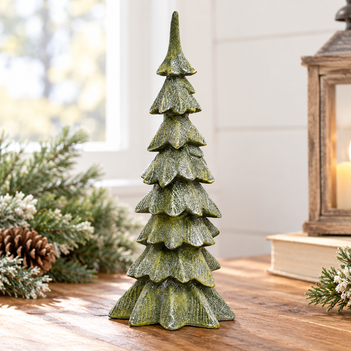 Resin Glittered Christmas Tree featuring a tall, slender evergreen silhouette with multiple irregular tiered branches, dimensional mossy green coloring, carved texture, and a shimmering silver glittered finish. The freestanding 10-inch resin tree is displayed on a rustic wood tabletop in a bright farmhouse-style holiday setting, accented by softly blurred frosted evergreen branches, pinecones, neutral books, and a warm lantern. Soft natural daylight highlights the tree’s textured surface and subtle sparkle 