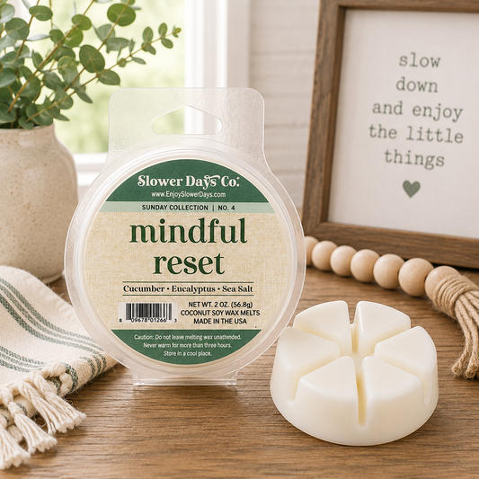 A round wax melt package labeled 'Slower Days Co. mindful reset' sits on a wooden surface next to a single white segmented wax melt.