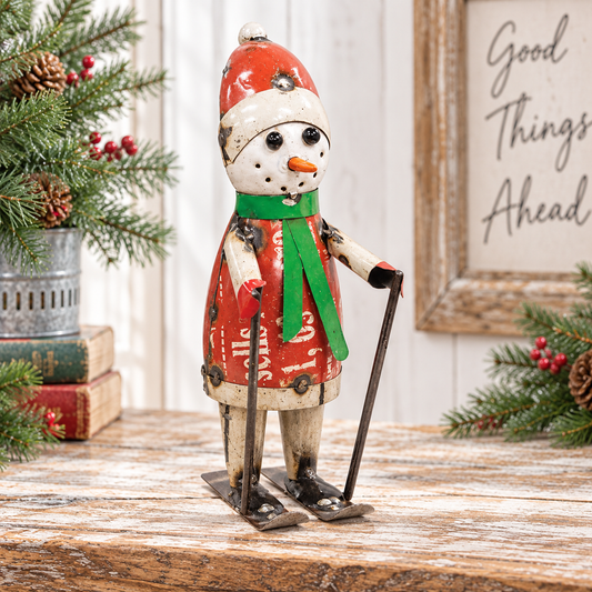A decorative snowman figurine dressed in a red coat and green scarf, holding ski poles and standing on skis on a rustic wooden surface. The scene is styled with pine branches, red berries, stacked books, and a framed sign reading 'Good Things Ahead'.