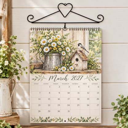 A spiral-bound March 2027 wall calendar hanging from a black metal hanger against a white wood-paneled wall. The calendar cover features a rustic floral illustration with daisies, a birdhouse, and a small bird.