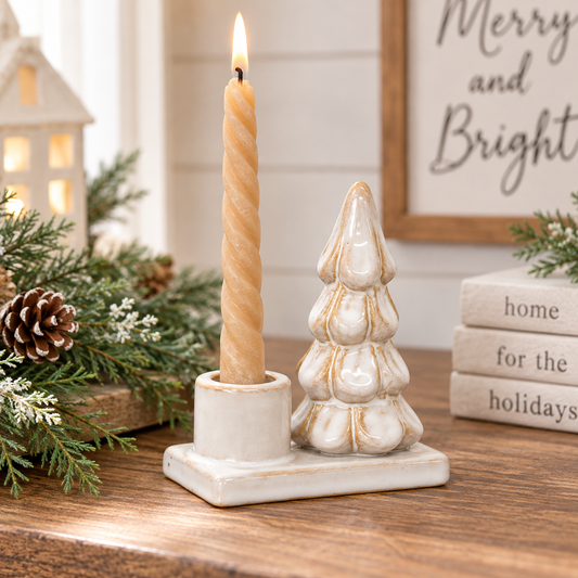 A ceramic holiday candle holder set on a wooden table, featuring a beige spiral taper candle in a cylindrical holder and a small sculpted Christmas tree candle holder next to it. The scene is decorated with pine branches, a pinecone, a small lit lantern, and a framed sign reading 'Merry and Bright' in the background.