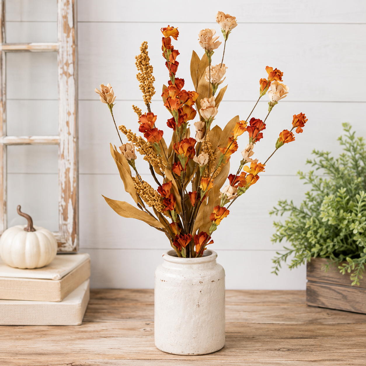 A faux autumn floral arrangement in a distressed white ceramic jar, styled on a wooden tabletop with a rustic white paneled wall background, a small white pumpkin, books, and a green plant.