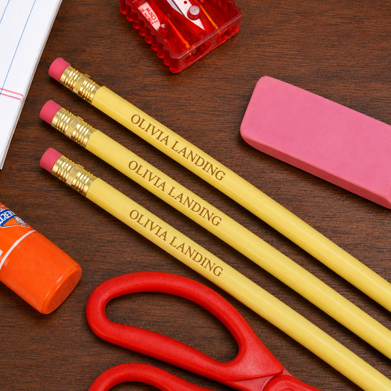 Three yellow pencils with pink erasers and gold custom text, arranged on a dark wood desk with a red sharpener, pink eraser, orange glue stick, and red scissors.