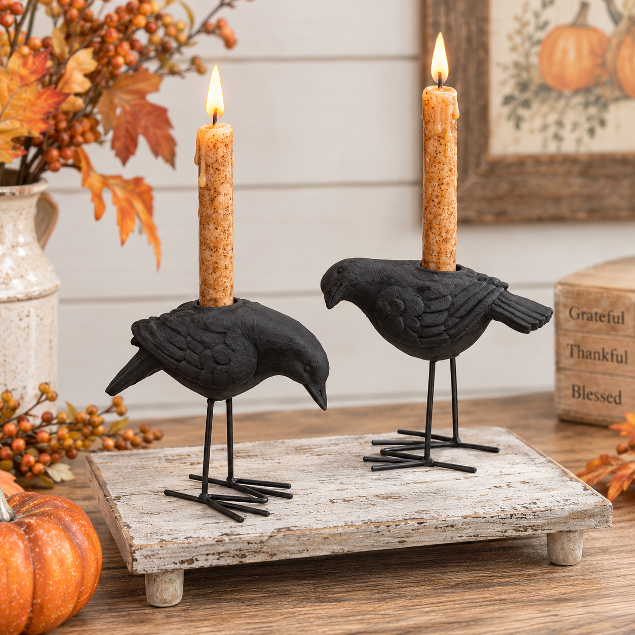 Two black bird candle holders shaped like crows or ravens, each holding a lit taper candle, displayed on a rustic white wooden board with autumn decor in the background.
