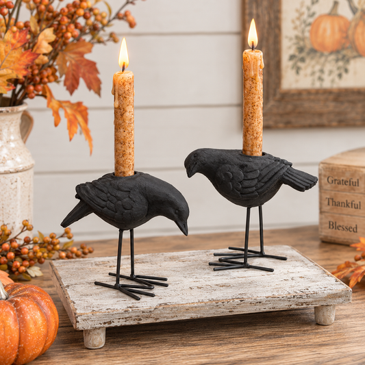 Two black bird candle holders shaped like crows or ravens, each holding a lit taper candle, displayed on a rustic white wooden board with autumn decor in the background.