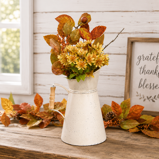 A rustic white speckled metal pitcher vase filled with yellow and orange autumn flowers, styled on a wooden table with fall leaves, a pinecone, and a framed sign against a white wood paneled wall.
