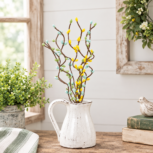 Decorative artificial willow branches with yellow and mint green buds in a distressed white ceramic pitcher, styled on a wooden table in a rustic room setting.