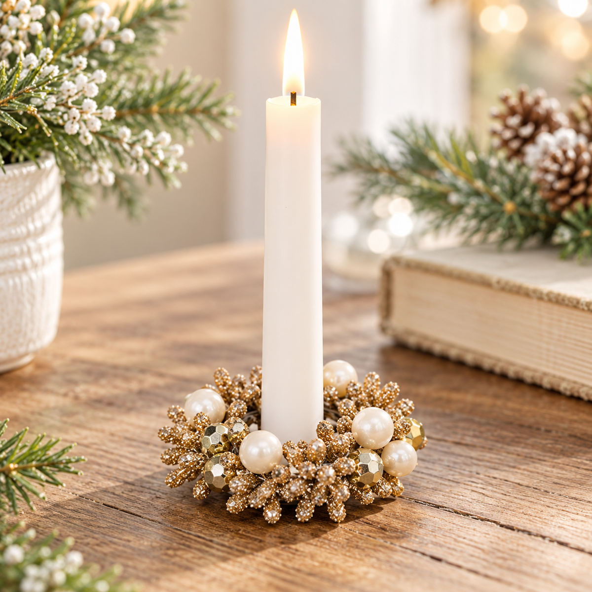 A lit white taper candle in a decorative gold and pearl holiday wreath, placed on a wooden tabletop with festive greenery and a book in the background.
