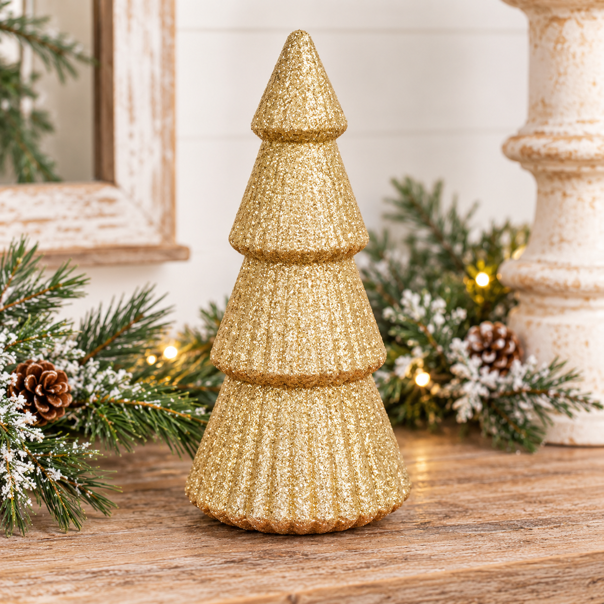 A small decorative Christmas tree in textured gold glitter stands on a wooden surface, surrounded by green pine branches, white snow-like dusting, pinecones, and warm string lights.
