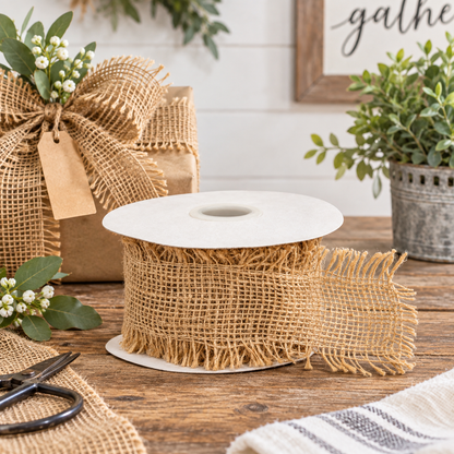 A spool of natural burlap ribbon with fringe edges, displayed on a rustic wooden table with a burlap-wrapped gift box and greenery in the background.