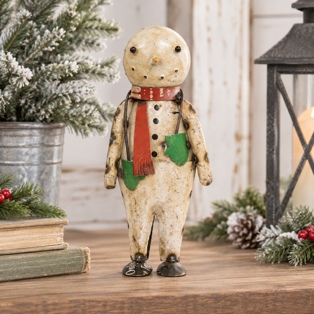 A rustic vintage-style snowman figurine standing on a wooden surface, decorated with a red scarf, green pockets, and black buttons, surrounded by holiday greenery and a lantern.