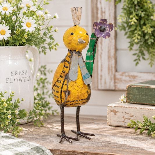 A rustic metal bird figurine with a yellow body, black wire legs, and a purple flower on a green stem, displayed on a wooden surface with flowers, greenery, and stacked books in the background.