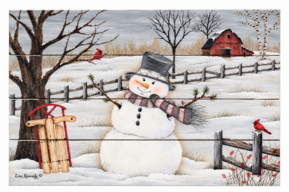 A winter landscape art print featuring a snowman in a snowy field with a red barn, bare trees, a fence, and red cardinals. The print has a horizontal wood-plank texture and is signed by artist Lisa Kennedy in the lower left corner.