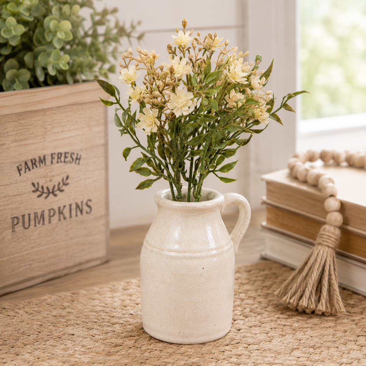 A small cream-colored ceramic pitcher vase filled with artificial cream flowers and green stems, styled on a woven rug next to a wooden crate and books.