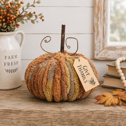 A decorative autumn pumpkin with vertical ribbed stripes in rust orange, mustard yellow, olive green, and taupe, featuring a rustic metal scroll stem and a 'Give Thanks' paper tag, displayed on a wooden table in a styled home decor setting.