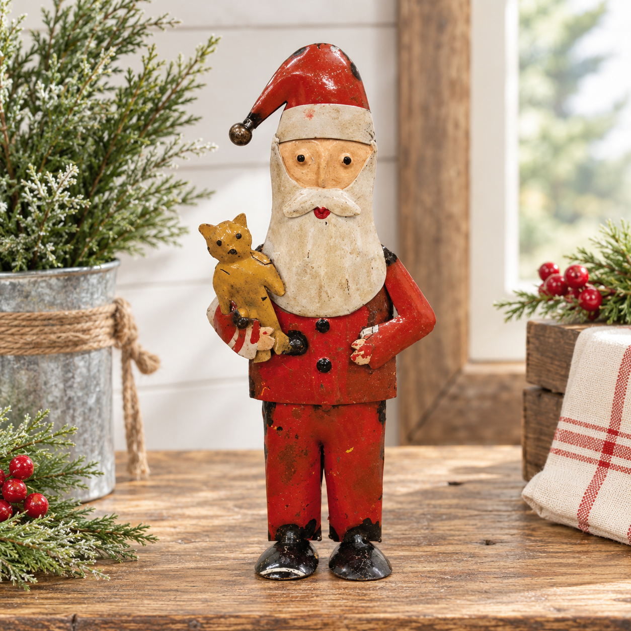 A rustic painted Santa Claus figurine holding a small golden dog, displayed on a wooden table with holiday greenery and a plaid cloth in the background.