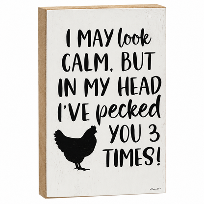 A rectangular wooden sign with a distressed white face and natural wood edges, featuring black text that reads 'I MAY look CALM, BUT IN MY HEAD I'VE pecked YOU 3 TIMES!' above a black chicken silhouette.