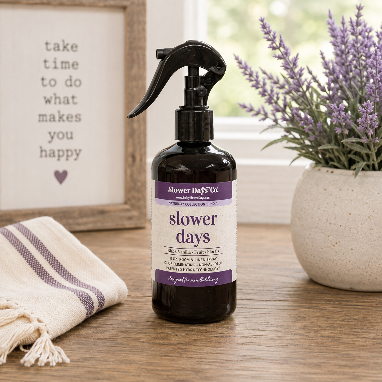 A dark amber spray bottle labeled 'slower days' by Slower Day Co., placed on a wooden table with a potted lavender plant and a framed sign in the background.