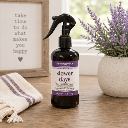 A dark amber spray bottle labeled 'slower days' by Slower Day Co., placed on a wooden table with a potted lavender plant and a framed sign in the background.