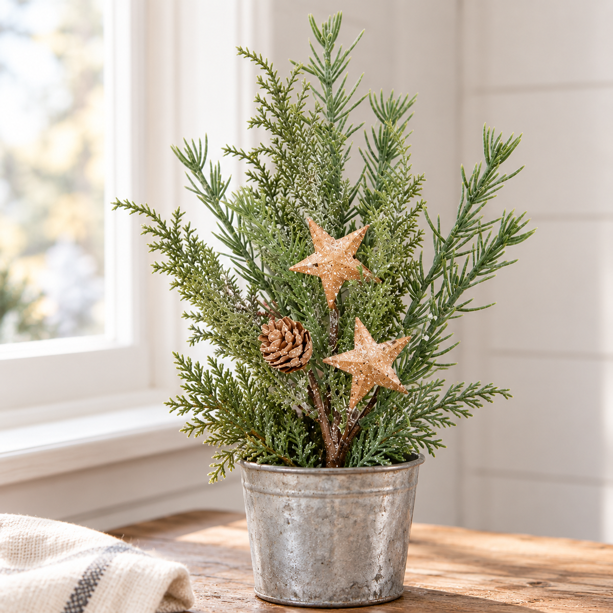 A small artificial evergreen tree arrangement in a galvanized metal bucket pot, decorated with two glittery gold stars and a pinecone, set on a wooden table near a window.