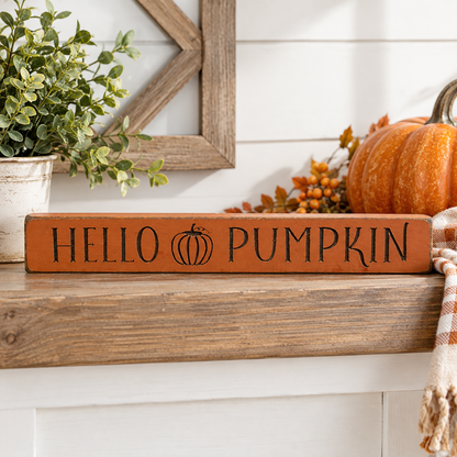 A rustic rectangular wooden sign with 'HELLO PUMPKIN' in black lettering and a small pumpkin icon, placed on a wooden mantel with fall decor including a pumpkin, plant, berries, and a plaid throw.