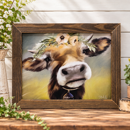 A framed rustic art print featuring a close-up painted cow with a flower crown, displayed on a wooden surface against a white wood plank wall with decorative plants around it.