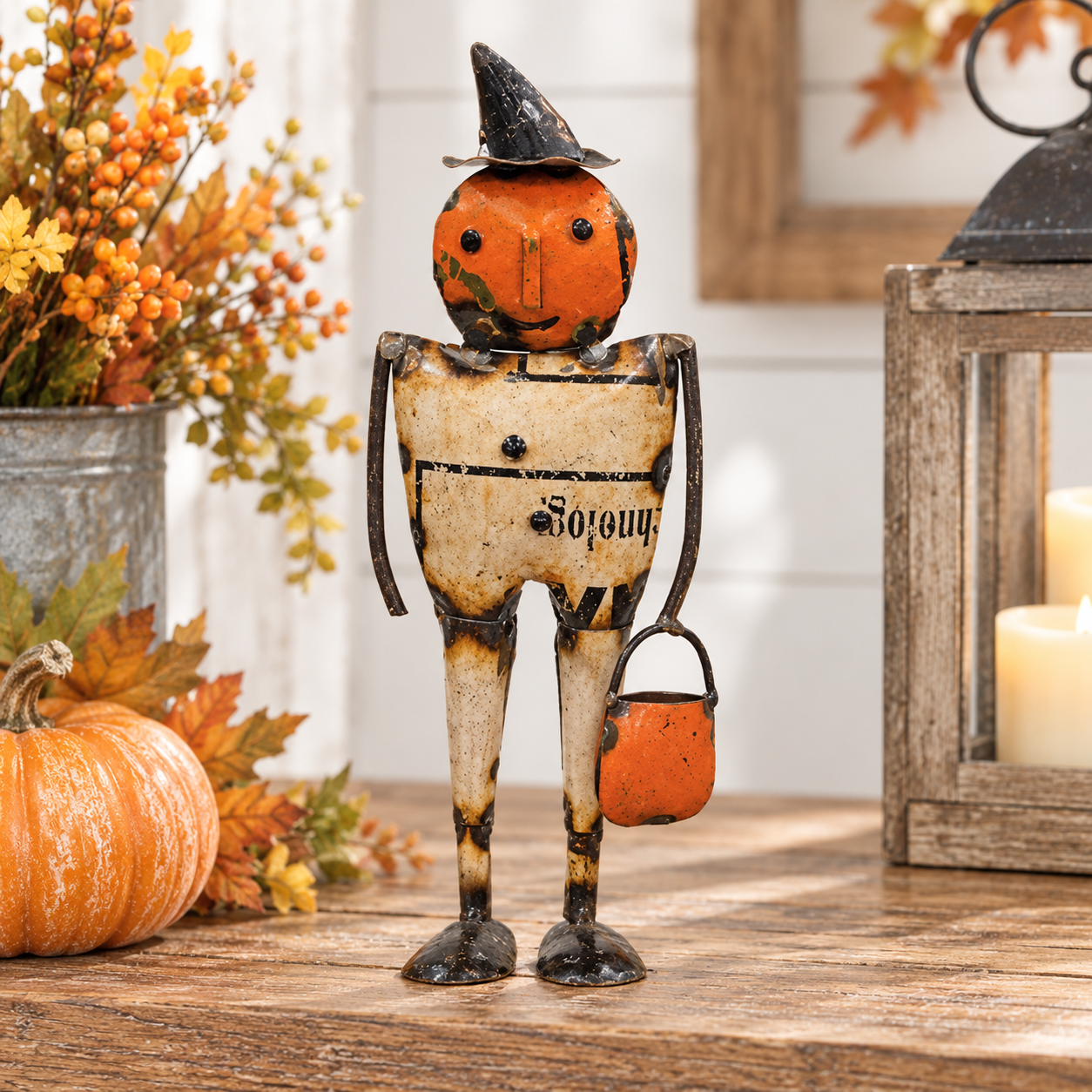 A rustic Halloween metal figurine shaped like a weathered figure with a pumpkin head, black hat, and orange bucket, displayed on a wooden surface with fall decor including a small pumpkin, artificial leaves, and candles.