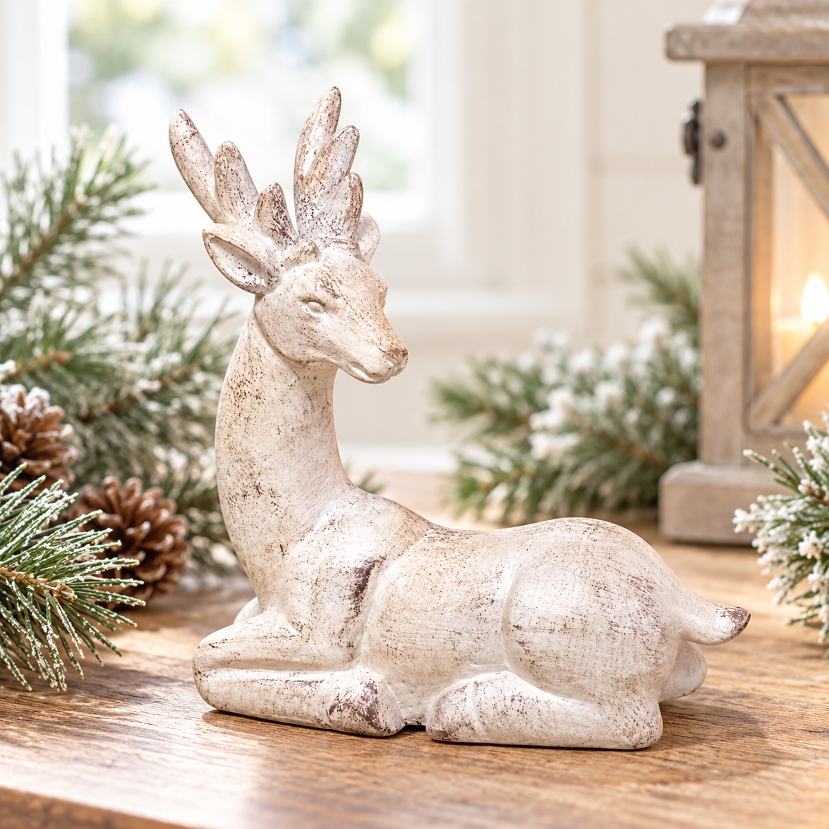 Distressed white resin reindeer figurine resting with its legs tucked beneath its body, featuring a long curved neck, side-turned head, detailed antlers, pointed ears, and a rustic whitewashed finish with natural-looking brown distressing. Styled on a warm wood tabletop with softly frosted evergreen branches and pinecones, with a wooden lantern and bright natural window light creating an airy farmhouse-style winter setting.