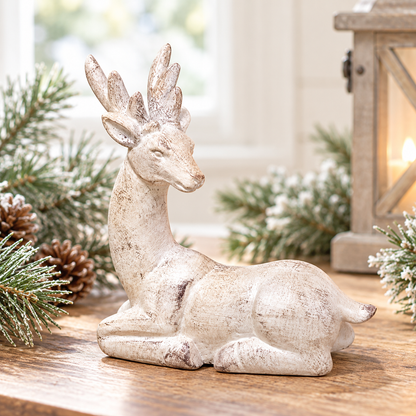 Distressed white resin reindeer figurine resting with its legs tucked beneath its body, featuring a long curved neck, side-turned head, detailed antlers, pointed ears, and a rustic whitewashed finish with natural-looking brown distressing. Styled on a warm wood tabletop with softly frosted evergreen branches and pinecones, with a wooden lantern and bright natural window light creating an airy farmhouse-style winter setting.