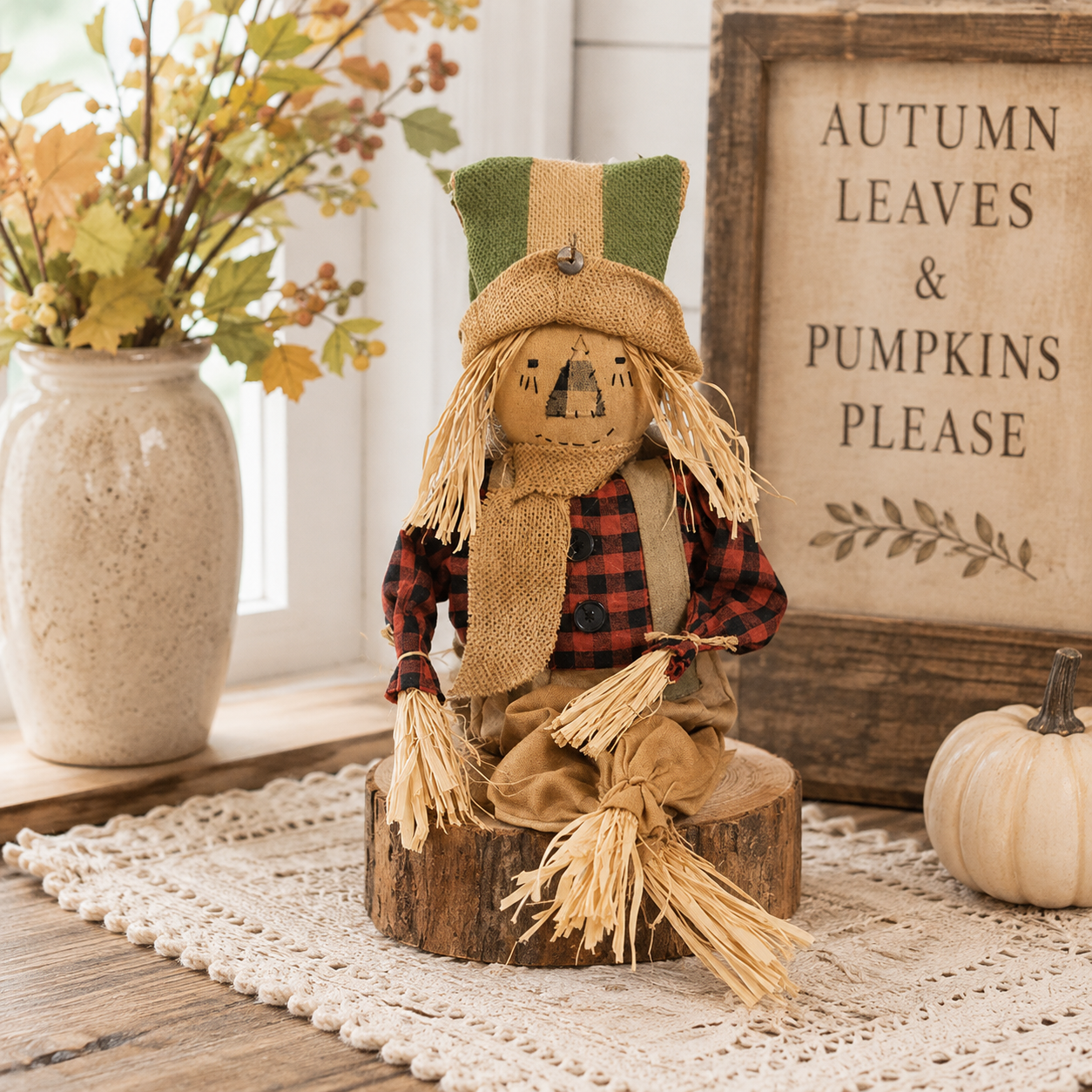 A rustic autumn scarecrow doll sits on a small wooden stump on a cream lace runner. The scarecrow wears a green and tan hat, a red and black buffalo plaid shirt, tan pants, and a burlap face covering.