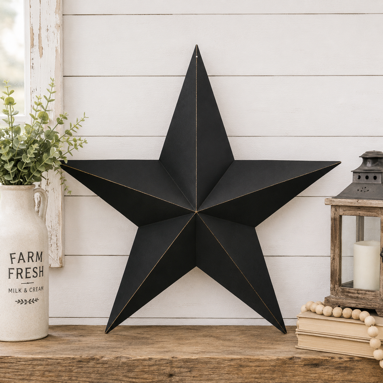 A large black metal star wall decor piece with gold-toned edge trim, displayed on a rustic wooden surface against a white shiplap wall. Decorative props include a white ceramic vase with greenery on the left and a lantern with books on the right.
