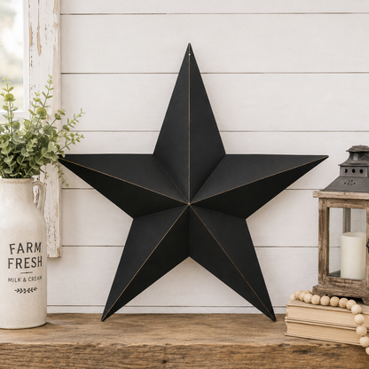 A large black metal star wall decor piece with gold-toned edge trim, displayed on a rustic wooden surface against a white shiplap wall. Decorative props include a white ceramic vase with greenery on the left and a lantern with books on the right.
