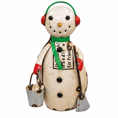 A vintage tin snowman holiday decoration with a weathered cream body, green scarf and ear muffs, red gloves, and black buttons, holding a small bucket and a shovel against a white background.