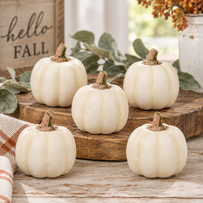 Five small faux white pumpkins arranged on a rustic wooden board and tabletop with fall decor, including greenery, a sign reading 'hello FALL,' and dried orange flowers.