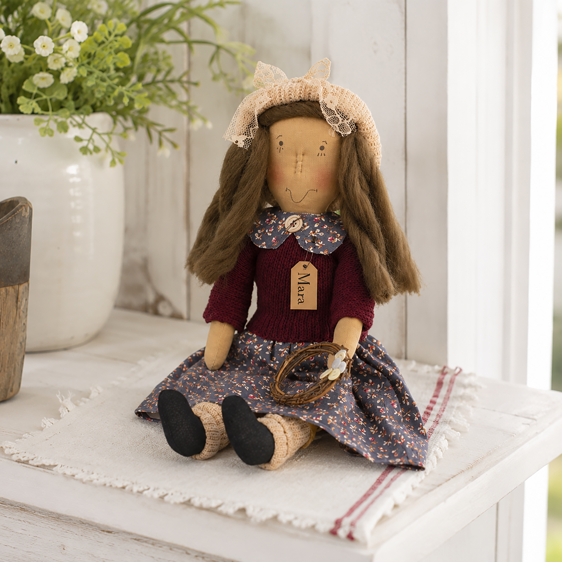 Mara plush shelf sitter doll with embroidered facial features, rosy cheeks, dusty blue floral dress, burgundy knit sweater, pale pink knit hat, and a rustic twig wreath with a white button flower, perfect for farmhouse, primitive, cottage, and country home décor.