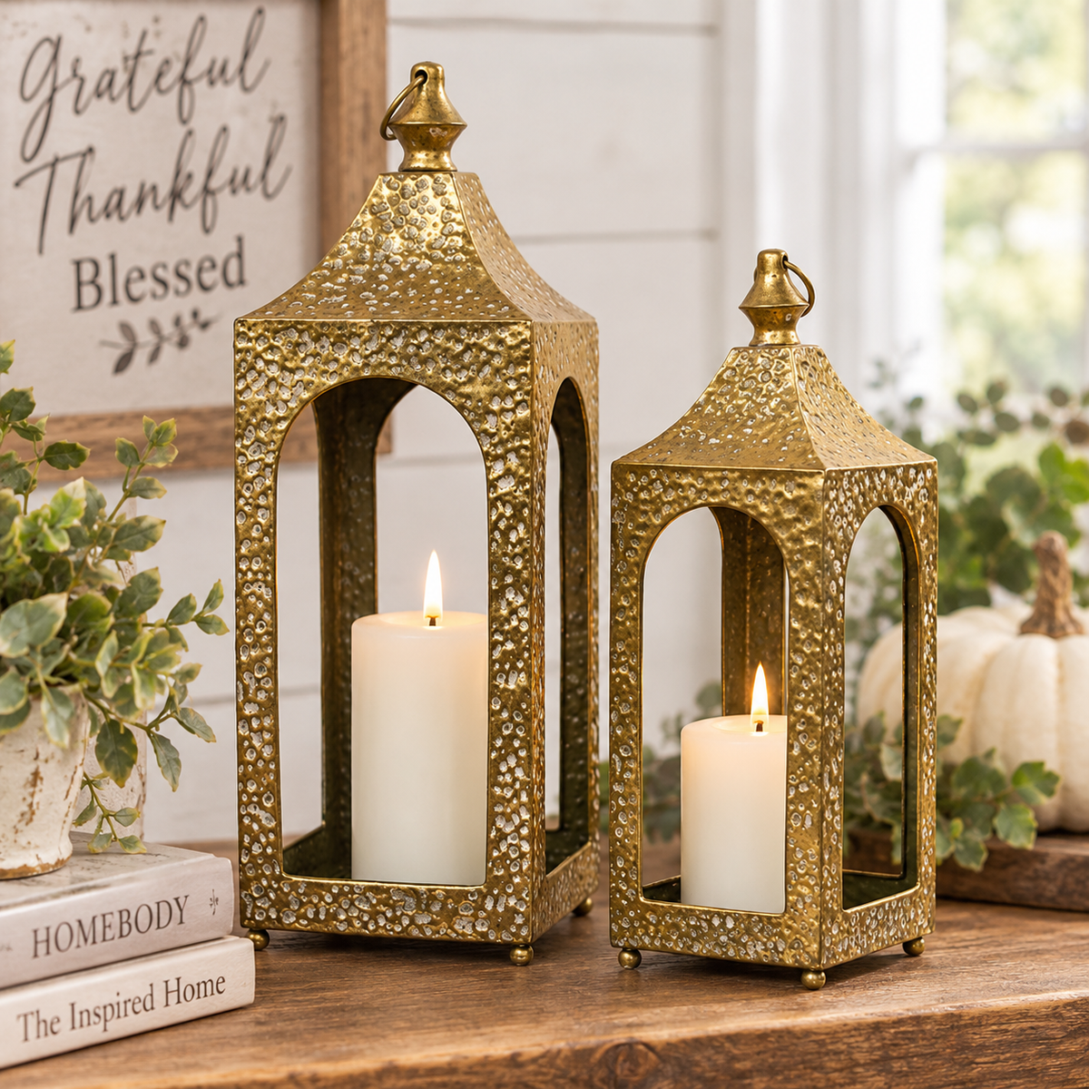 Two decorative gold-toned metal lanterns with textured hammered finishes, each holding a white pillar candle, styled on a wooden surface with pumpkins, greenery, and a framed sign in the background.