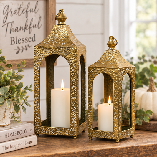 Two decorative gold-toned metal lanterns with textured hammered finishes, each holding a white pillar candle, styled on a wooden surface with pumpkins, greenery, and a framed sign in the background.